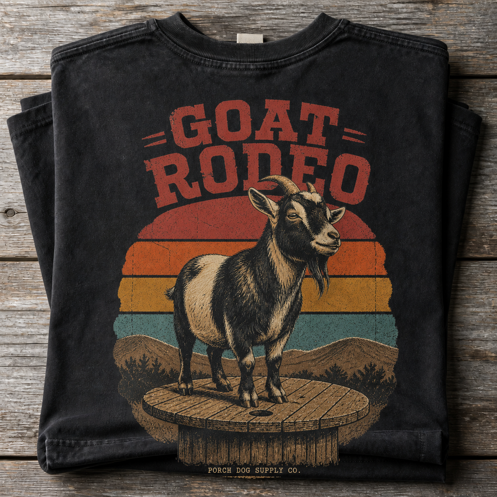 Goat Rodeo vintage-washed black tee folded on weathered barnwood, back art of a pygmy goat on a cable spool